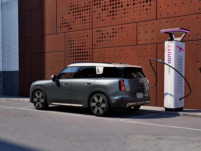 Vue latérale d’un MINI Countryman stationné dans une rue de la ville pendant qu’il se recharge à une borne de recharge électrique IONITY à côté d’un mur perforé de couleur rouille.